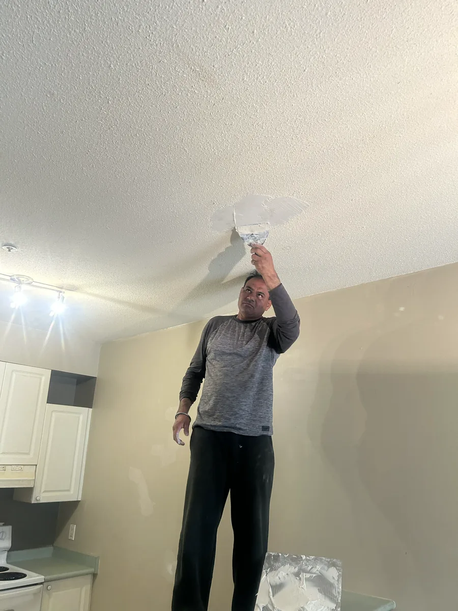 A renovation worker preparing a textured ceiling with a scraper.